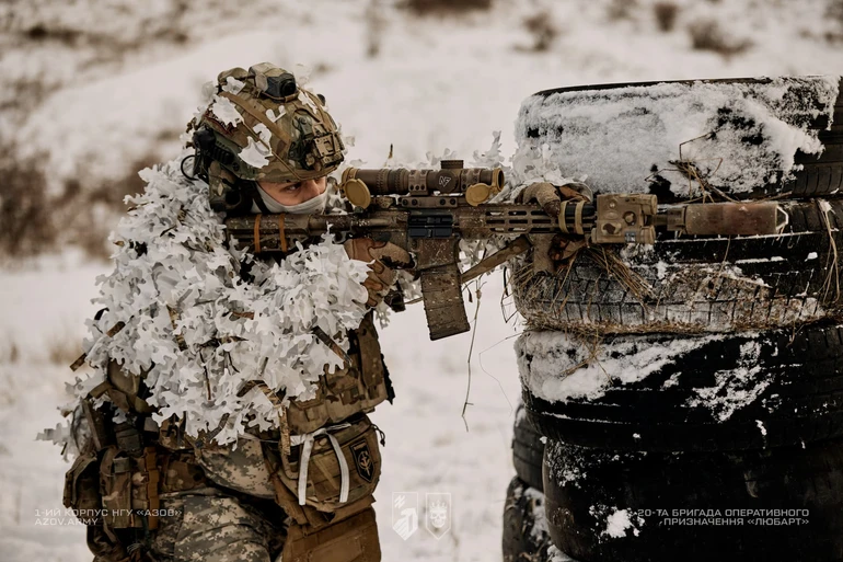 Боєць у зимовому маскувальному спорядженні веде прицільний вогонь з укриття з автомобільних шин під час тренування на засніженому полігоні.
