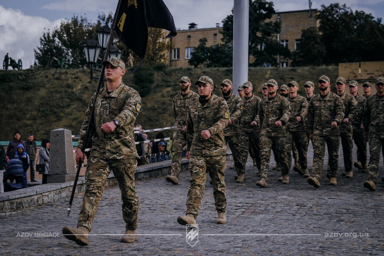 Колона військових у камуфляжі марширує містом з прапором.