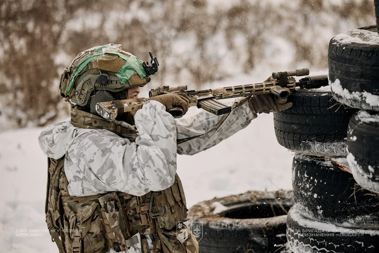 Військовий у білому камуфляжі стріляє з автомата, спираючись на імпровізоване укриття з шин у зимових умовах.